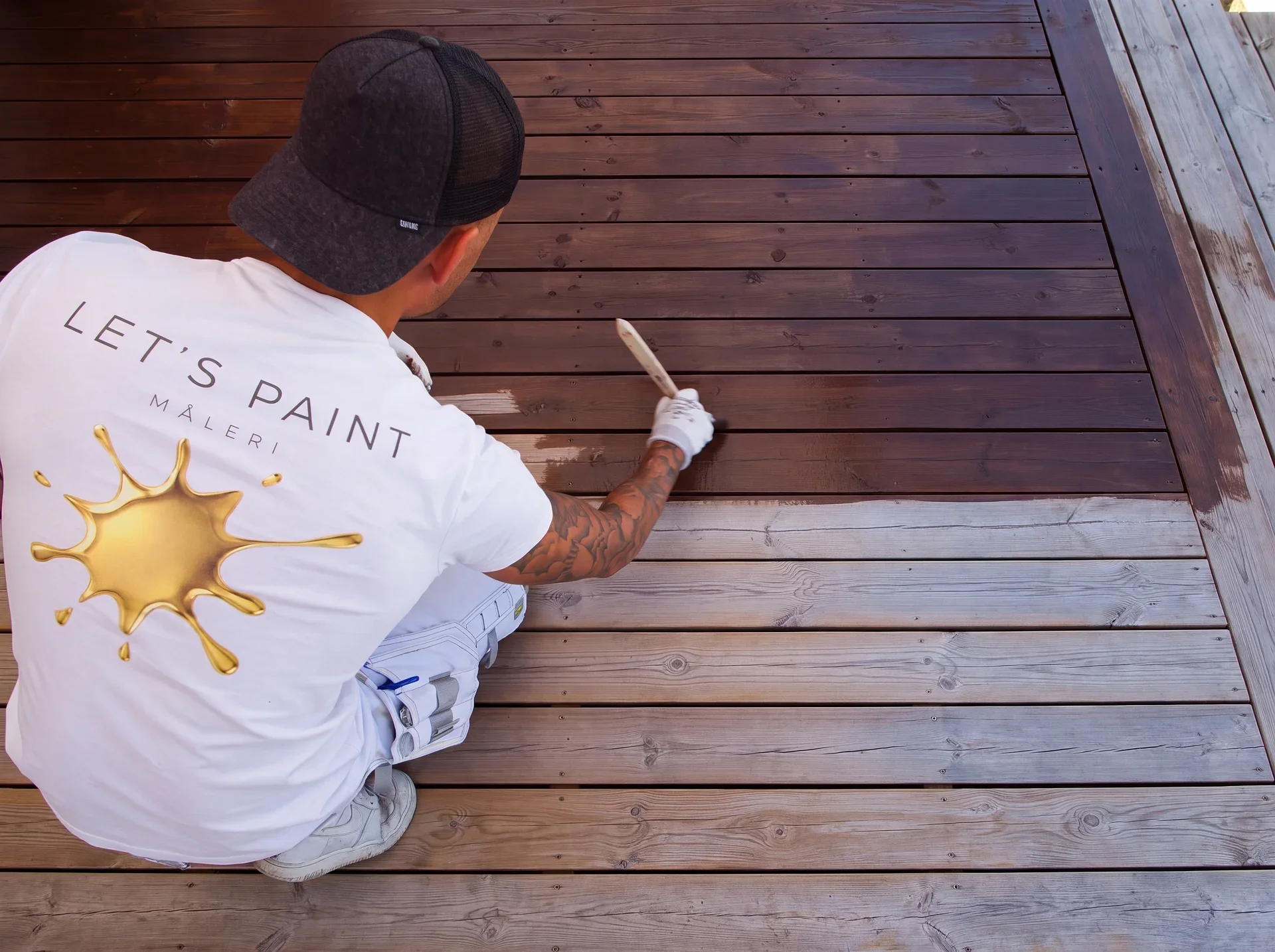Man målar trädäck med träolja, bär "Let's Paint"-tröja och keps.